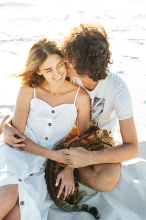 Cheerful young couple having fun on the beach with their bengal cat