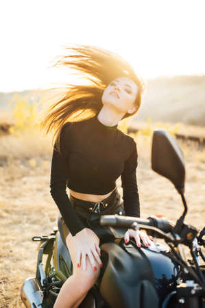 Beautiful girl in black clothes sits on motorcycle and poses for camera. Evening sunsetの写真素材