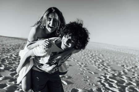 Beautiful couple having fun on the beach. Cheerful girl jumped on her boyfriend backの写真素材