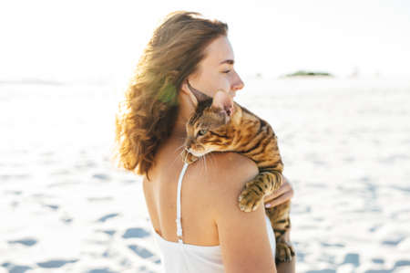 Cute girl in white dress on the beach hugs her bengal catの写真素材