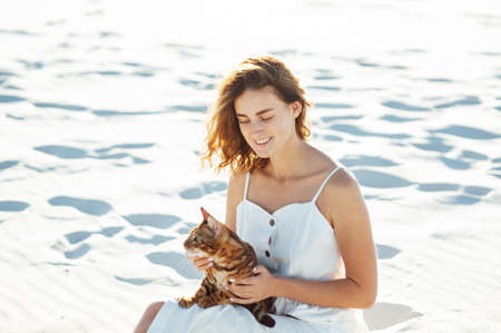 Cute girl in white dress on the beach hugs her bengal catの写真素材