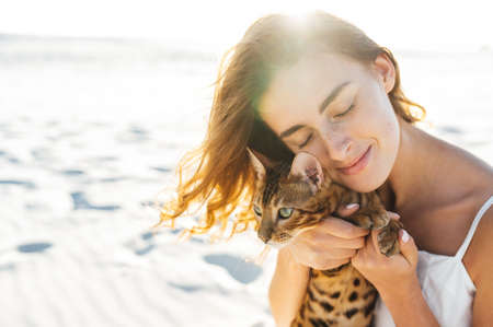 Cute girl in white dress on the beach hugs her bengal cat