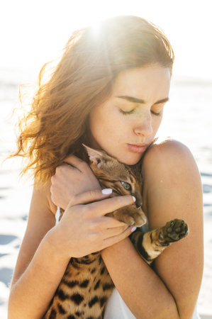 Cute girl in white dress on the beach hugs her bengal catの写真素材