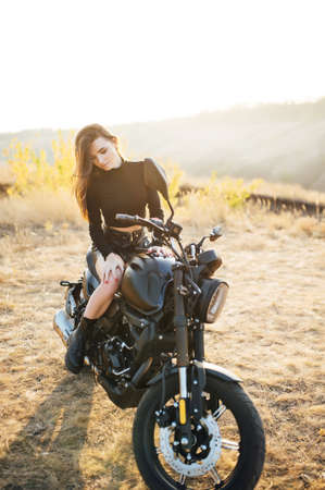 Beautiful girl in black clothes sits on motorcycle and poses for camera. Evening sunsetの写真素材