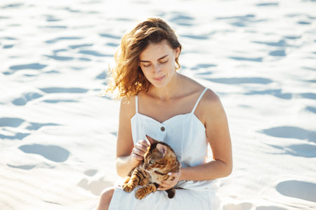 Cute girl in white dress on the beach hugs her bengal catの写真素材
