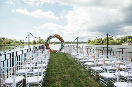 Round arch for a wedding ceremony by the river. White glass chairs stand for guestsの写真素材