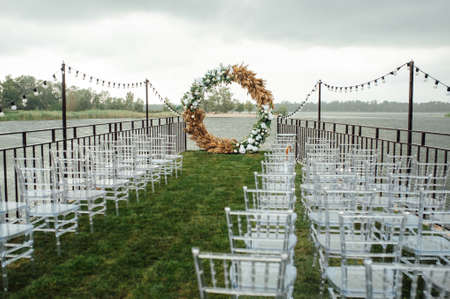 Round arch for a wedding ceremony by the river. White glass chairs stand for guestsの写真素材