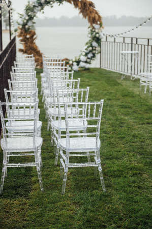 White glass chairs stand on the green grass. Round arch for a wedding ceremony In the background . Raining dayの写真素材