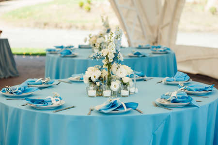 Wedding tables for guests. Round tables with a blue tablecloth. Decorated with flowers, plates, glasses and moreの写真素材