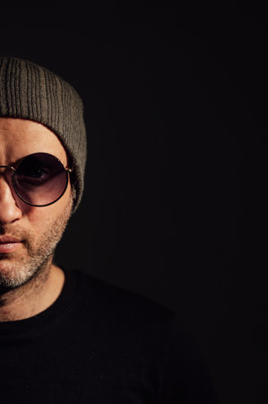 Serious dramatic man in sunglasses looking away while standing against black studio backgroundの写真素材