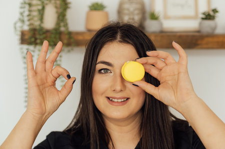Smiling woman holding tasty colored macaron covering her eyesの写真素材