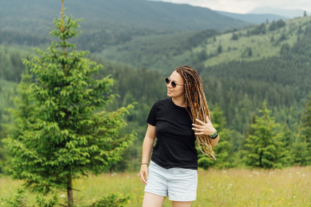 A beautiful woman with a hairstyle - a lot of braids, posing at the top of the mountain in a black T -shirt and shorts.の写真素材