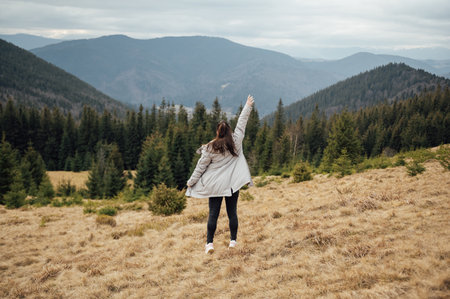 Happy girl stands in the mountains, enjoys the beautiful view and raises her hand upの写真素材