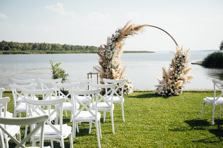 Round arch for a wedding ceremony by the river. White glass chairs stand for guestsの写真素材