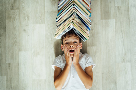 A cheerful boy lies on the floor, and above his head there is a large stack of books in the form of a roof.の写真素材