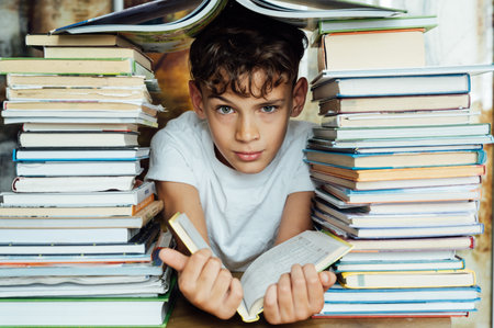 A cheerful boy looks through two stacks of books. On the head is a book in the shape of a roofの写真素材