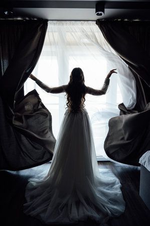 The girl stands with her back near the window and opens the curtains with her handsの写真素材