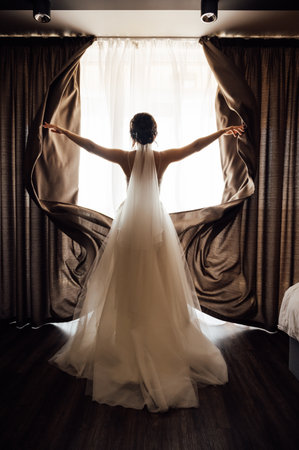 The girl stands with her back near the window and opens the curtains with her handsの写真素材