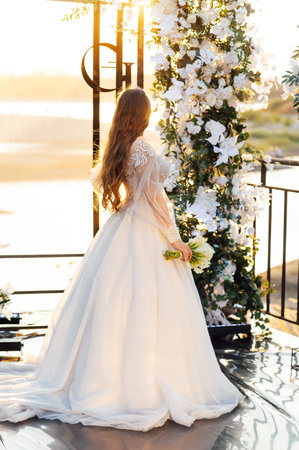 A brunette bride in a white dress holds a bouquet and walks to the wedding arch, among many white chairs. Wedding ceremony in nature. Celebrationの写真素材