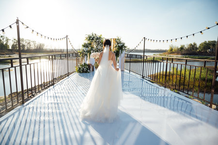 A brunette bride in a white dress holds a bouquet and walks to the wedding arch, among many white chairs. Wedding ceremony in nature. Celebrationの写真素材