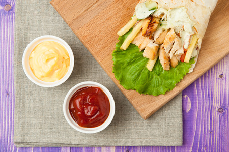 Arabic shawarma on wooden board and two white cups with ketchup and cheese on purple wooden tableの写真素材