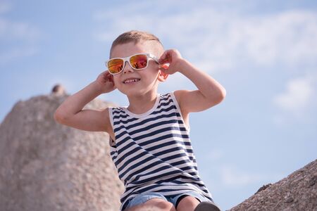 Little caucasian boy in a vest and sunglasses siting on breakwater block on the background of the sky with cloudsの写真素材