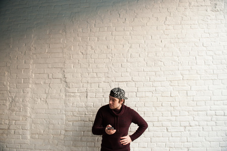 Handsome young man in baseball cap and sweater with phone in hands on white brick wall background with copyspaceの写真素材