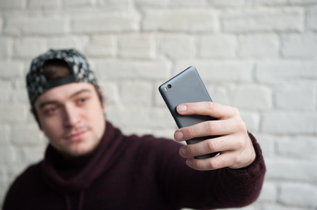 cute young man in a cap and sweater makes selfie with smartphone on brick wall backgroundの写真素材