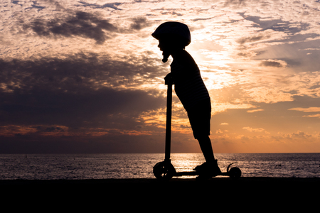 Silhouette of a little boy wearing helmet standing on scooter against the background of a sea sunsetの写真素材