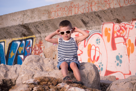 Beautiful little boy in trendy sunglasses and sailor vest sitting on stone in front of wall with graffitiの写真素材
