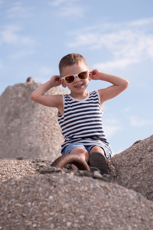 Cute smiling little boy in sunglasses and singlet sitting on a concrete breakwaterの写真素材