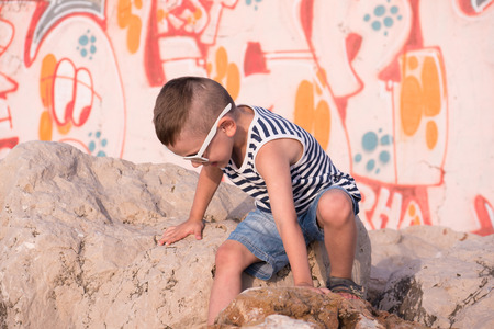 little boy wearing sunglasses and shorts climbing the rocks in front of graffiti wallの写真素材