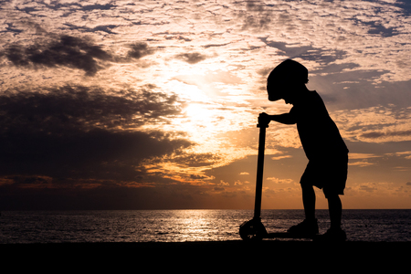Silhouette of sporty little boy standing on scooter against background of sea sunsetの写真素材