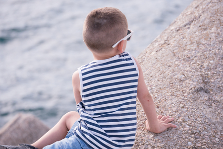 healthy little boy dressed in a striped shirt and jean shorts sitting on concrete breakwater by the sea looking afarの写真素材