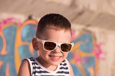 smiling small kid wearing sunglasses with sea sunset reflection in it and sailor shirt on graffiti background in summertimeの写真素材