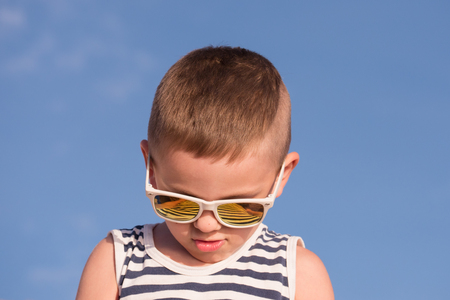 smiling little boy wearing white sunglasses with stripes reflections and sailor stripes singlet on blue sky backgroundの写真素材