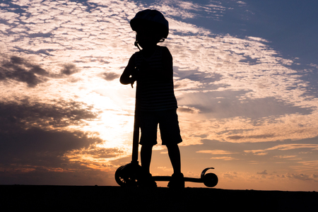 Silhouette of small boy wearing helmet with scooter on background of beautiful evening sky with cloudsの写真素材