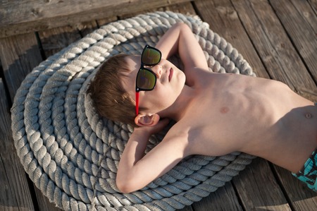 beautiful little boy in sunglasses relaxes lying on the rope and the wooden pierの写真素材