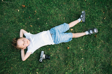 handsome little boy lying on the green grass with fallen leaves near film cameraの写真素材