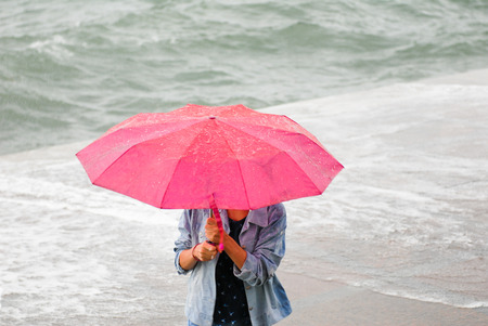 woman with red umbrella escaping from the rain and hurricane on sea coast background in autumnの写真素材