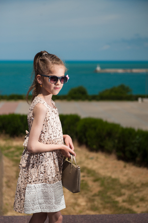 beautiful small girl wearing sunglasses and trendy dress holding bag with sea and shore backgroundの写真素材