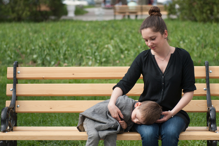 mother sitting on the bench gently embracing her small son sleeping on her hips holding tenderly her fingerの写真素材