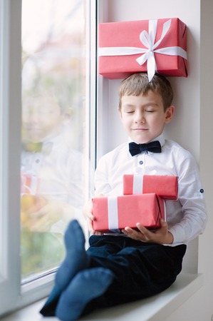 happy little boy in bow tie sitting by window with a gift box on his head and two gift boxes in hands closing his eyesの写真素材