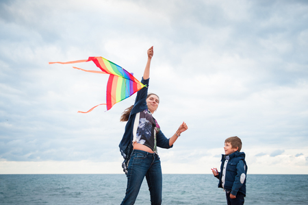 playful mother and child with flying kite playing on sea and cloudy sky background in autumnの写真素材