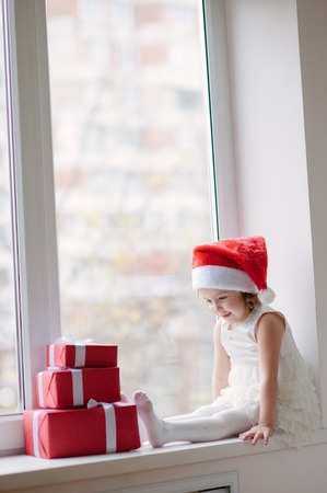 pretty little girl in Santa hat sitting at the window with three boxes giftsの写真素材
