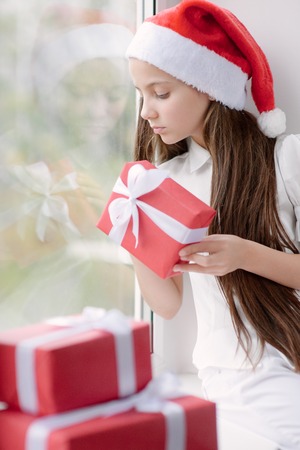 adorable little girl in Santa hat sitting by the window holding a box with gifts in Christmas Eveの写真素材