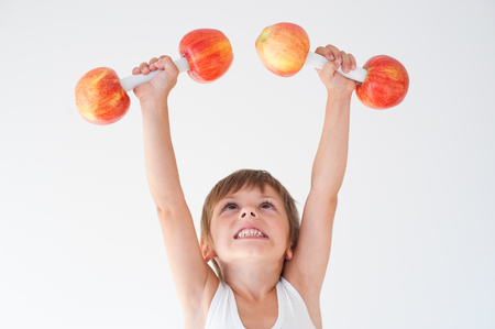 Cheerful cute kid lifting dumbbells made from red and yellow apples overheadの写真素材