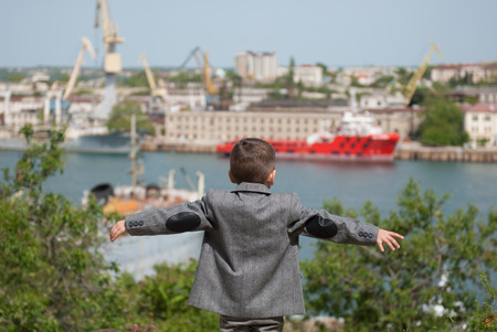 small boy in a jacket stands against the backdrop of a seaport, placing his hands in the sides imitating a flightの写真素材