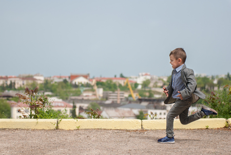 healthy little boy in a jacket running along the asphalt along the cityscape backgroundの写真素材