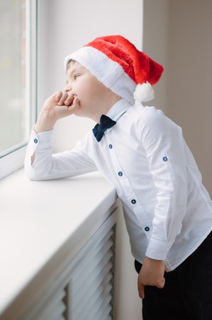 pensive boy in santa hat and bow tie at the window in Christmas Eveの写真素材